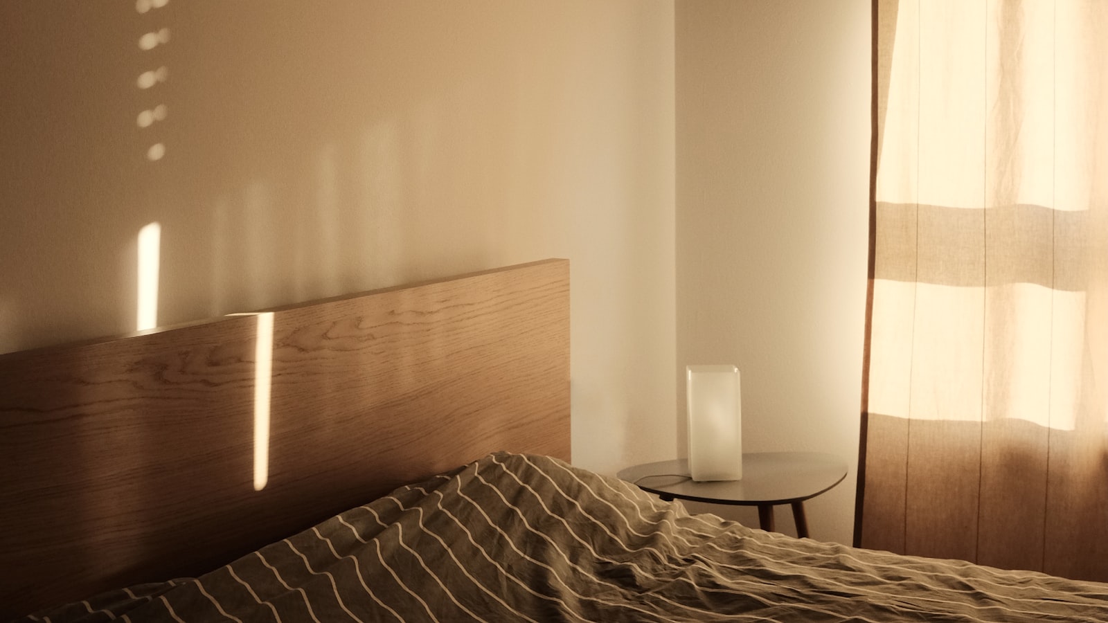 Afternoon sunlight falling through a linen curtain across a bed with striped bedding, a wooden headboard and a small lamp on a bedside table