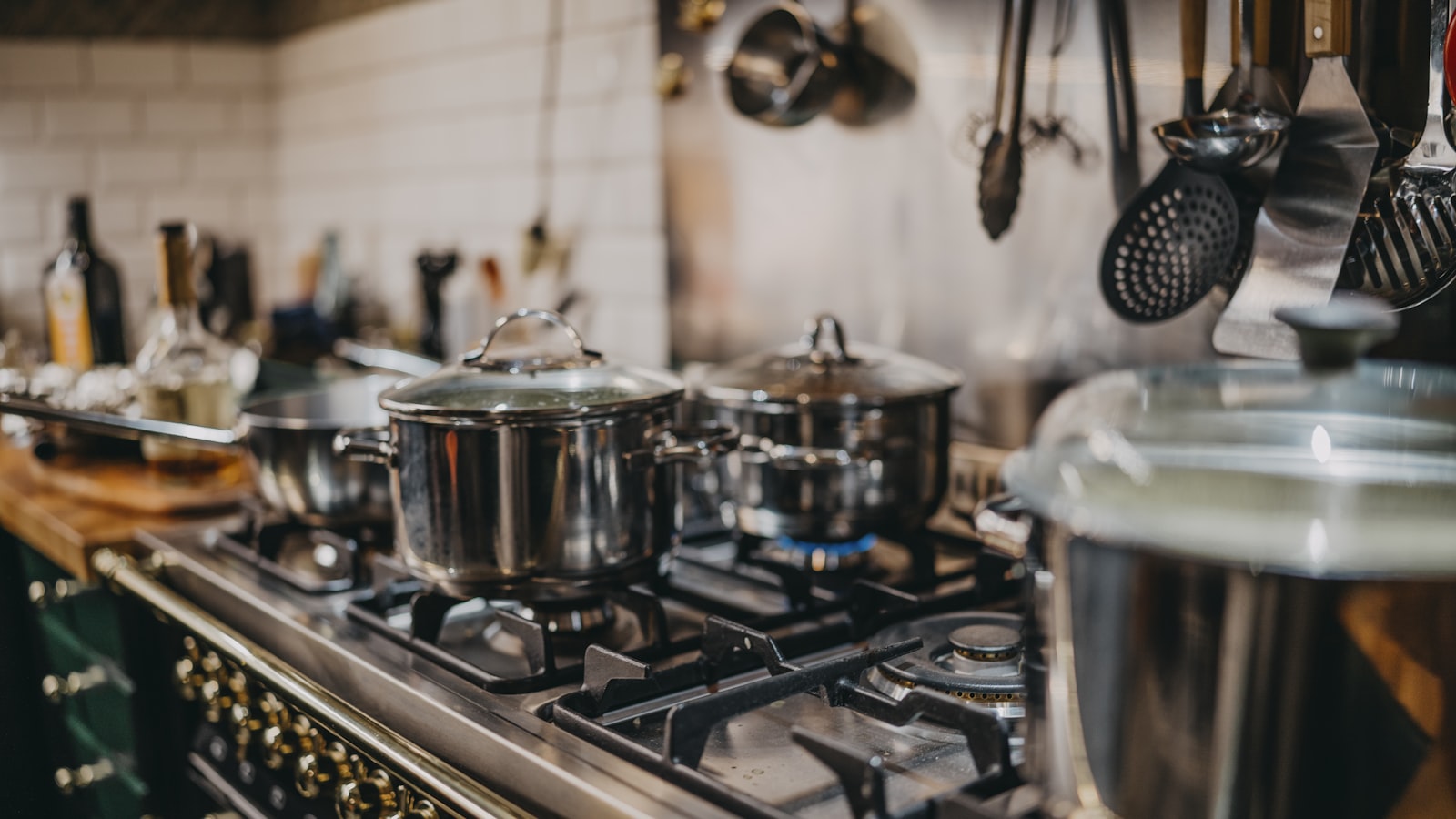 Stainless steel pans simmering on a gas hob in a home kitchen, with utensils hanging above and white tiles behind