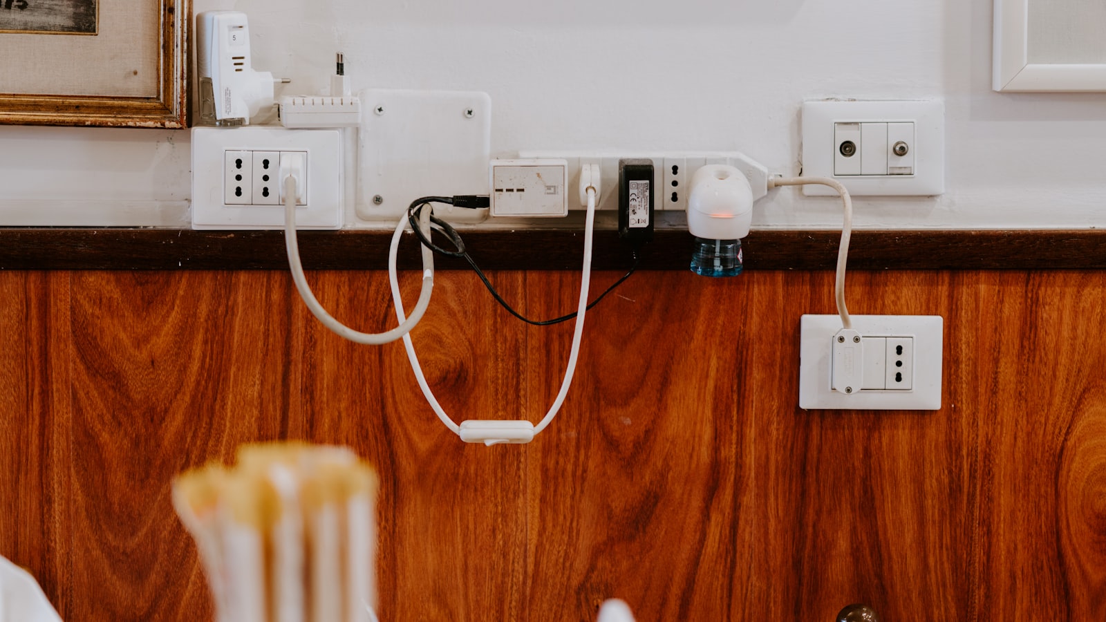 A row of white wall sockets above dark wood panelling, with several adapters, cables and a plug-in air freshener left connected