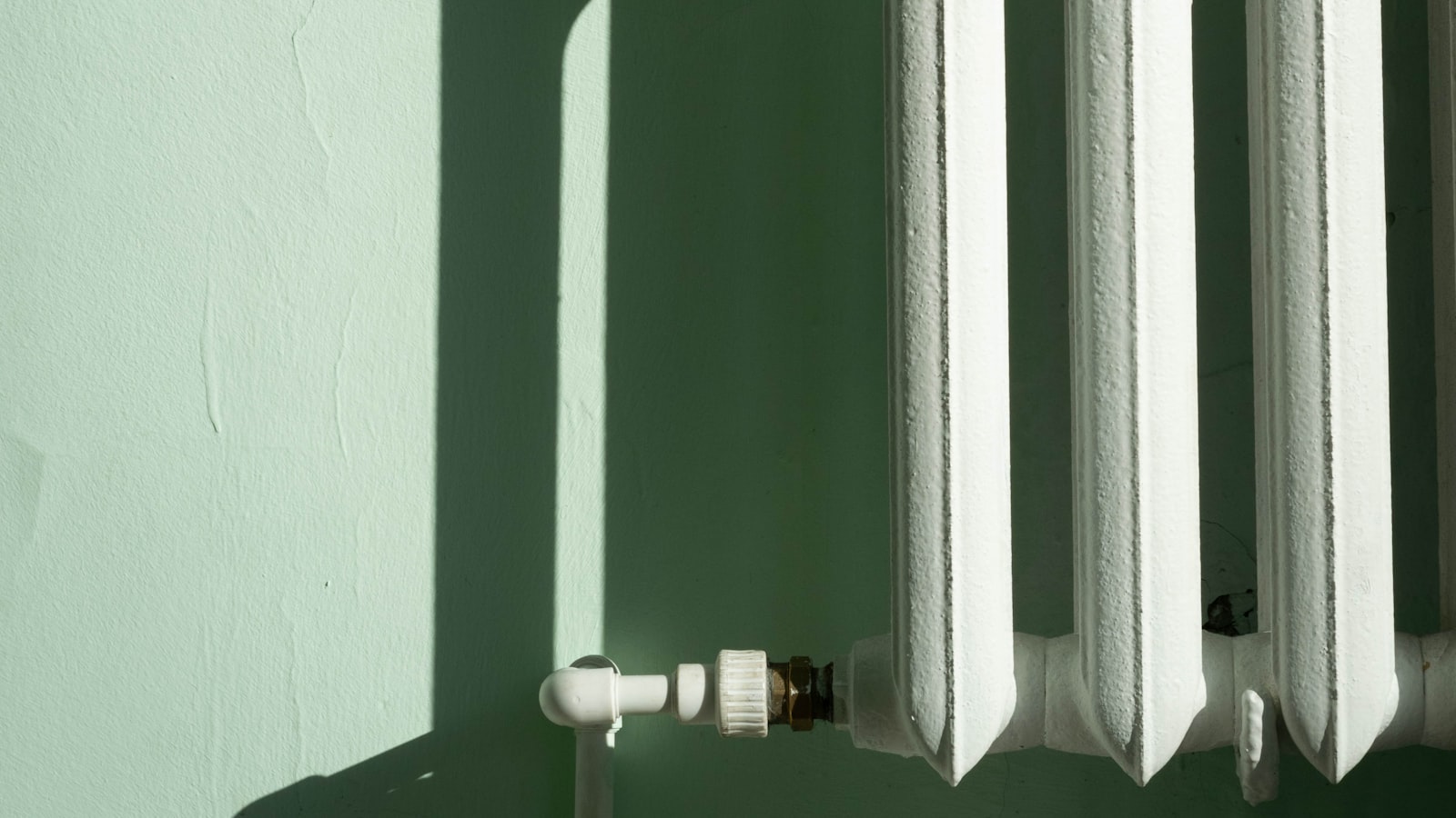 A white column radiator and its valve against a pale green wall, lit by hard diagonal sunlight