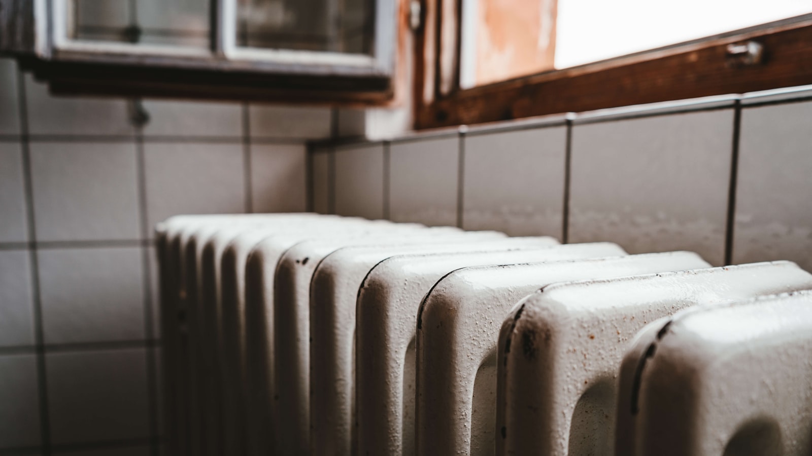 An old white cast iron radiator below a window in a tiled room, lit from the side by daylight