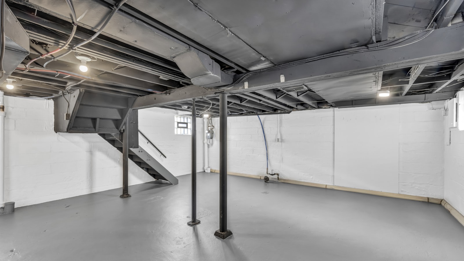 An empty unfinished basement with white painted block walls, steel support posts, exposed ceiling ducting and a staircase