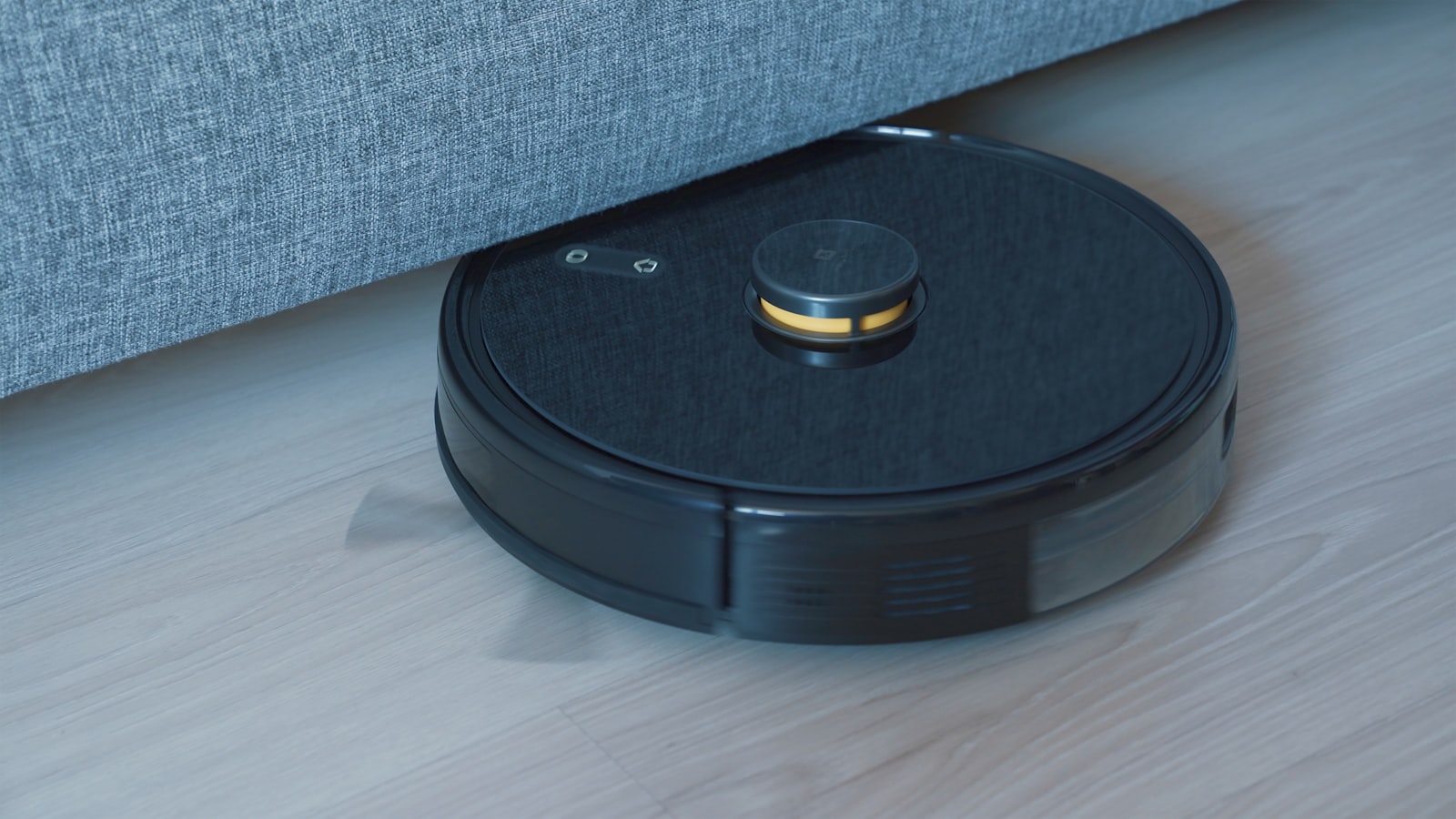 A black robot vacuum reversing out from under a grey sofa on a wooden floor