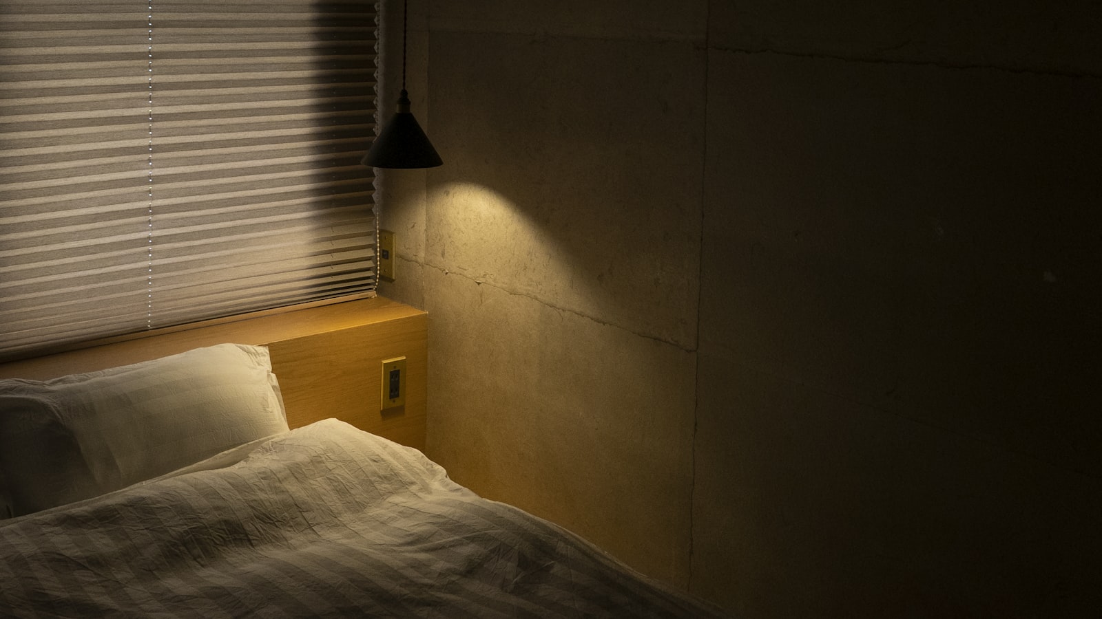 A bed with white pillows and a rumpled duvet in a dim bedroom, lit by a small pendant lamp hanging beside closed window blinds