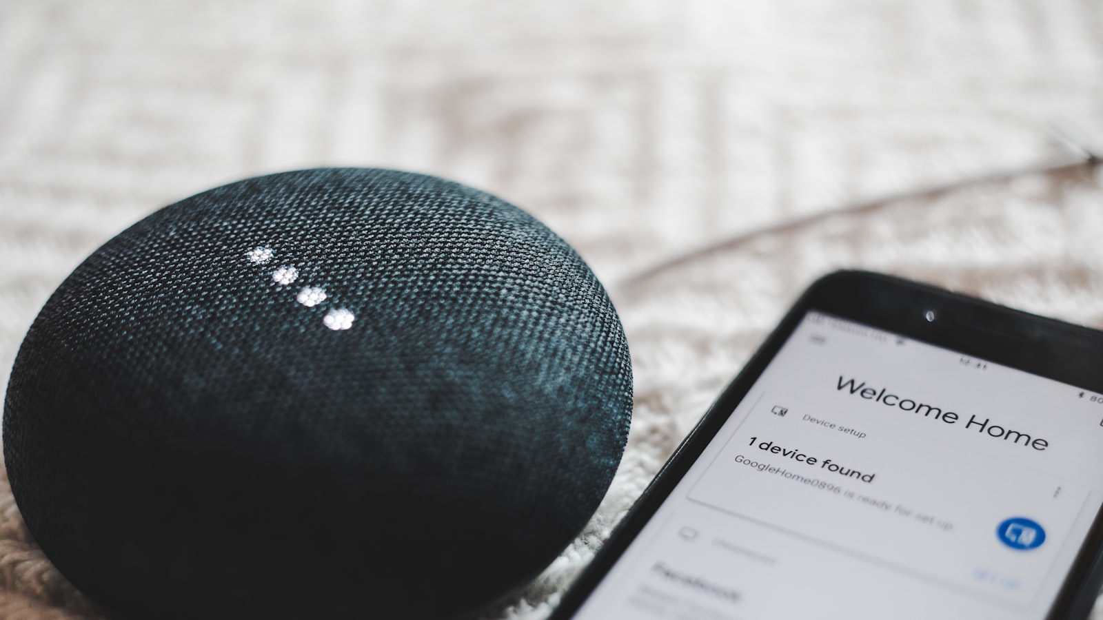 A Google Home Mini speaker beside a phone showing a device-setup screen